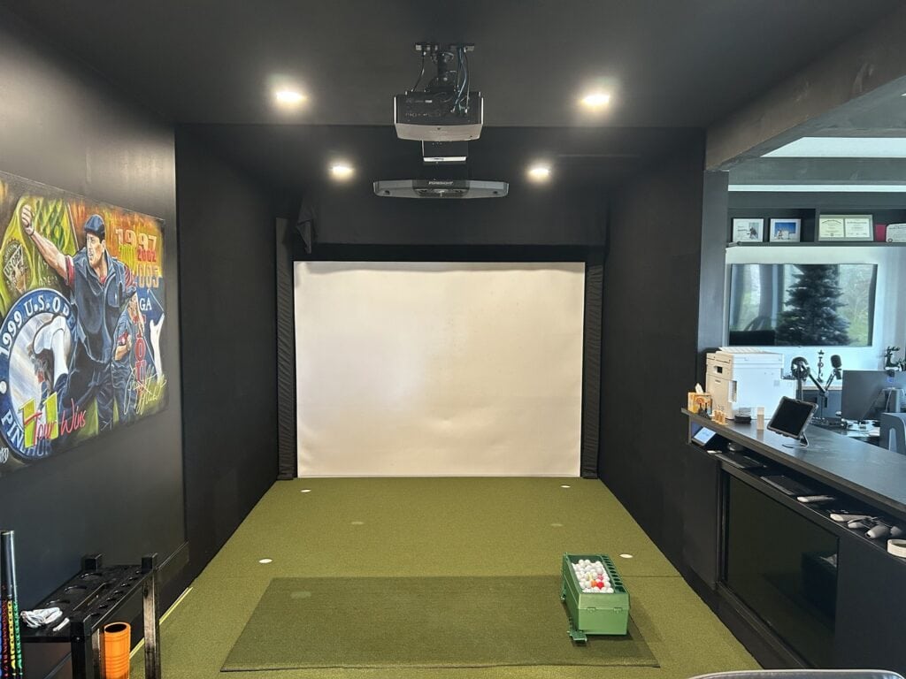 Interior view of a dedicated entertainment room featuring a golf simulator setup with a large impact screen and lounge seating.