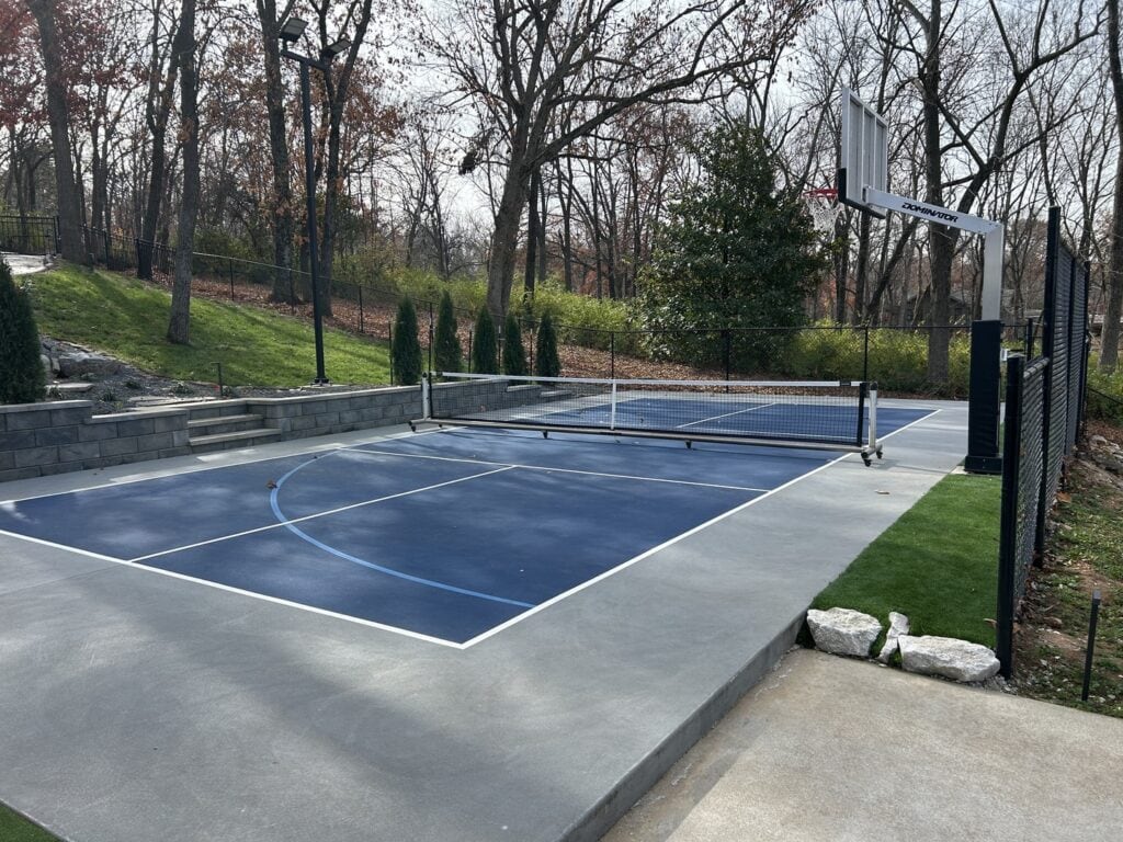 An outdoor multi-purpose sports court set up for pickleball and basketball, surrounded by black chain-link fencing.